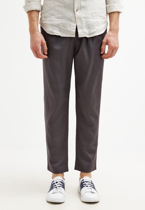 Mann trägt hochgekrempeltes hellbeiges Hemd, dunkelgraue gerade Hose und weiße Sneakers mit marineblauen Details, steht vor neutralem Hintergrund.