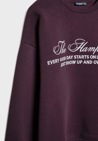 Burgundský sveter vyrobený z mäkkej tkaniny, s okrúhlym výstrihom. Biele vyšitie s nápisom "The Hamptons" a motivačnou frázou nižšie.
