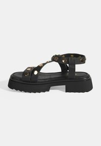 Black leather sandal with wide straps featuring gold studs, a thick ridged sole, and an open back design for easy wear.