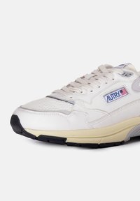 Sneaker atletico bianco realizzato con materiali in rete e pelle, dotato di una suola beige, dettagli grigi e un patch logo blu con dettagli rossi.
