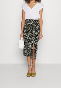 Černá midi sukně s bílým květinovým vzorem, boční rozparek, kombinováno s bílým tričkem s výstřihem do V, zelenými platformovými sandály a malou bílou kabelkou.