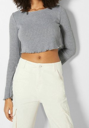 Femme vêtue d'un haut court gris côtelé à manches longues et d'un pantalon taille haute couleur écrue, debout devant un fond uni.