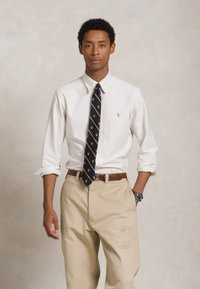 Biely pruhovaný košeľový top, modrá kravata so zlatým vzorom, béžové nohavice s jemnou výšivkou, hnedý opasok a hodinky na zápästí.