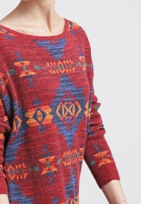 Roter, gestrickter Pullover mit bunten geometrischen Mustern in Blau, Orange und Grün. Verfügt über einen weiten Halsausschnitt und lange Ärmel.