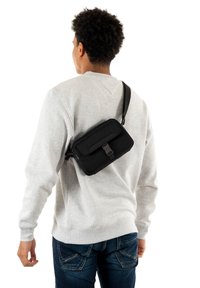 Borsa a tracolla nera con patta frontale, tracolla regolabile, forma rettangolare e texture opaca, indossata sopra un maglione grigio.