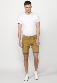 Rumene denim kratke hlače s zavihanimi hlačnicami, v kombinaciji z belo majico s kratkimi rokavi. Model nosi bele športne čevlje in stoji pred nevtralno ozadje.