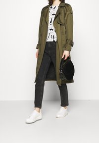 Trench coat verde oliva, top grafico nero con design astratto, jeans neri, sneaker bianche e borsa a tracolla nera. Texture morbide.
