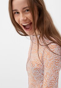 Langärmliges Oberteil aus weichem, elastischem Material mit einem wirbelnden orange-weißen Muster. Das Modell lächelt und hat mittelbraune Haare.