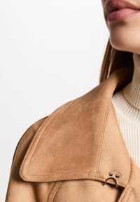 Veste en daim marron avec un grand col, ornée d'une touche de matériel doré et d'un col roulé crème à côtes en dessous. Texture lisse visible.