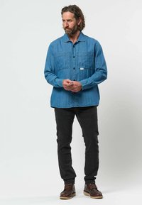 Homme aux cheveux bouclés mi-longs et à la barbe, porte une chemise en jean bleue, un jean noir et des bottes brunes, debout sur un fond uni clair.