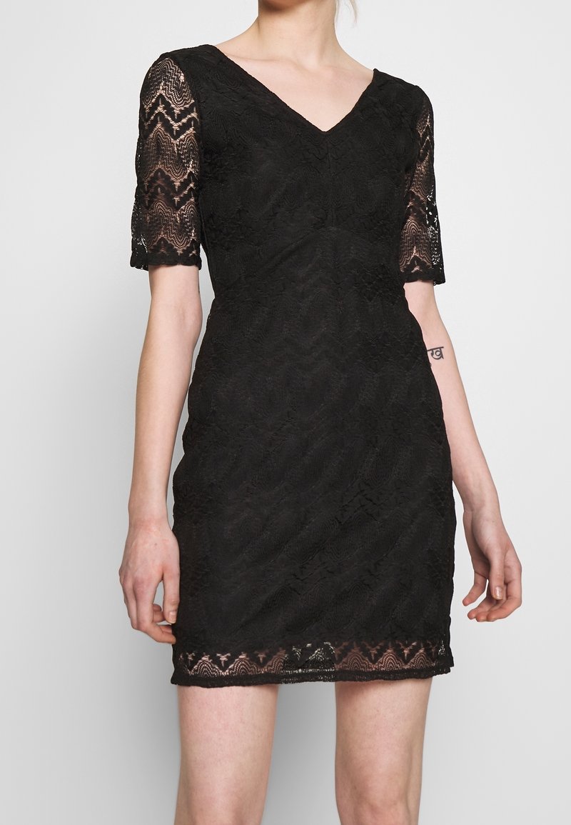 Femme portant une courte robe noire en dentelle avec un décolleté en V et des manches trois-quarts, montrant un petit tatouage sur son avant-bras intérieur.