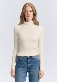 Pulover cu guler înalt, de culoare crem, cu model ribbed și mâneci lungi. Asortat cu blugi albastri cu talie înaltă, având cinci buzunare și detalii cu nasturi din cupru.
