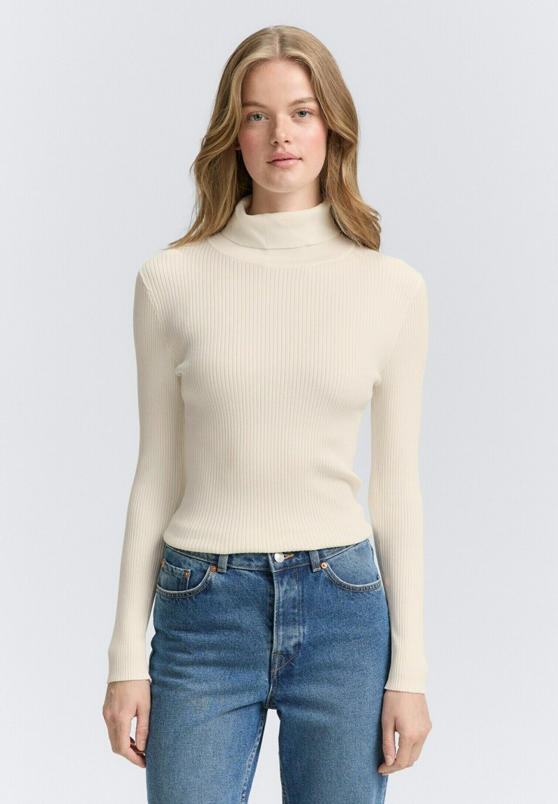 Pulover cu guler înalt, de culoare crem, cu model ribbed și mâneci lungi. Asortat cu blugi albastri cu talie înaltă, având cinci buzunare și detalii cu nasturi din cupru.