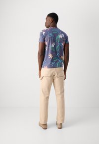 Camisa de manga corta en azul con estampado floral colorido, combinada con pantalones cargo beige. Material de algodón, corte holgado, detalle visible en el bolsillo trasero.