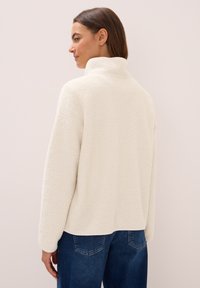 Femme aux cheveux bruns portant une veste en polaire blanche et un jean bleu, se tenant de côté contre un mur clair uni.