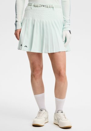 Kvinde iført en lyseblå plisseret nederdel med bælte, hvid langærmet top, golfhandske, hvide sokker og hvide sneakers.