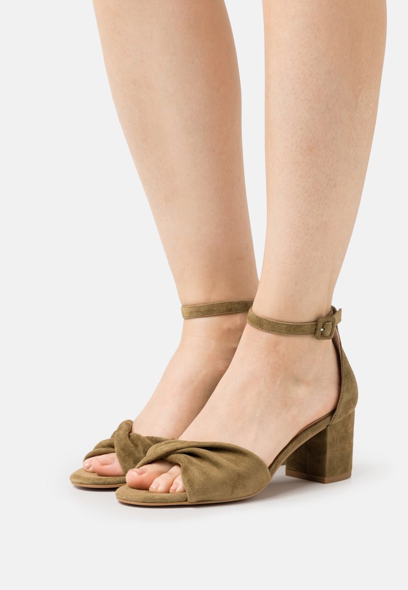 Pieds portant des sandales à talons carrés en daim vert olive avec des brides torsadées et des boucles à la cheville sur fond blanc.