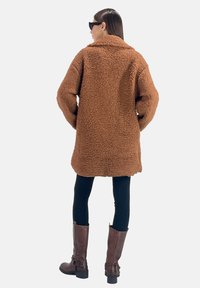 Manteau en shearling marron, oversized et duveteux, avec un col cranté, porté sur des leggings noirs et des bottines en cuir marron.