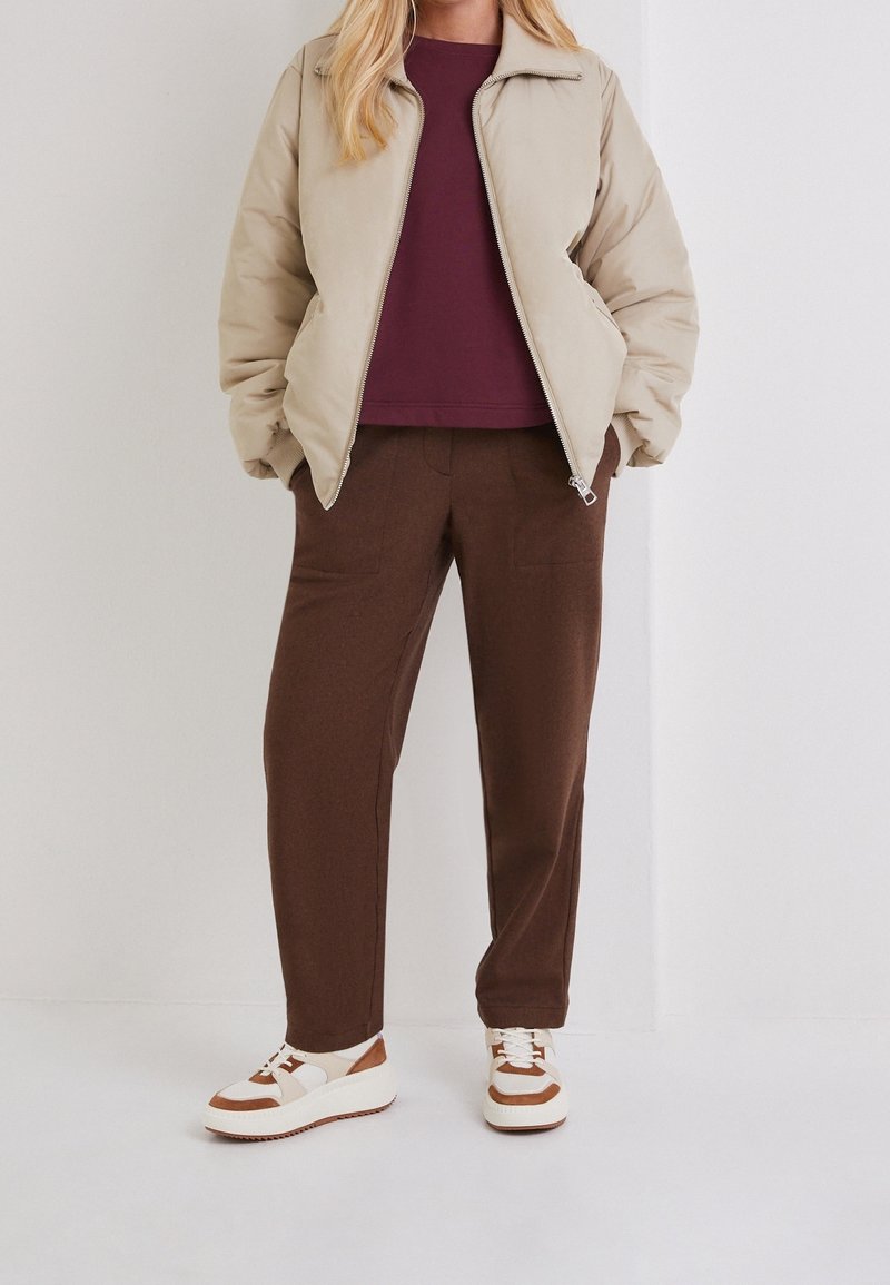 Chaqueta bomber beige con cremallera, sudadera burdeos, pantalones marrones y zapatillas blancas con detalles marrones. Conjunto casual de corte relajado y tejidos texturizados.