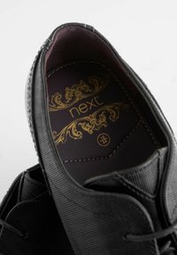 Zapato negro con una parte superior texturizada y un interior suave. La plantilla presenta letras doradas que dicen "siguiente" y detalles ornamentales sobre un fondo marrón oscuro.