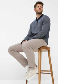 Dunkelgrauer Strickpullover für Herren mit navyfarbener Kragen, beigen Hosen und hellen Turnschuhen, sitzt auf einem Holzschemel.