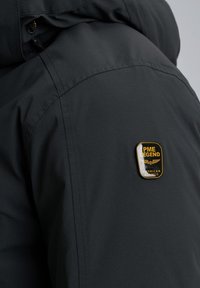 Zwarte synthetische jasstof met een subtiele ribbelstructuur. Bevat een gouden en zwarte PME Legend-embleem op de schouder.