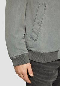 Szara kurtka bomberka o gładkiej teksturze, ze ściągaczami na rękawach i boczną kieszenią. Model nosi ciemne dżinsy, ręka oparta na kurtce.