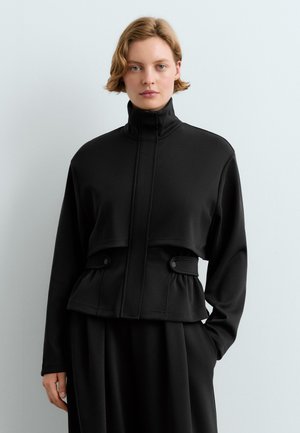 Frau mit kurzem Haar, die eine schwarze, hochgeschlossene Jacke mit geknöpften Taschen und einem gerafften Taillenbereich trägt, steht vor einem einfarbigen hellen Hintergrund.