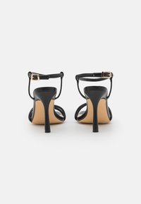 Sandalias de tacón alto de cuero negro con correa de tobillo ajustable y hebilla grabada, que presentan un diseño elegante y punta abierta.