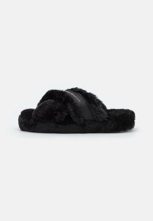 Sandale slide en peluche noire avec des brides croisées et une bande textile à logo sur le dessus, conçue pour le confort intérieur et extérieur.