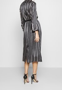 Vestido negro con patrones circulares rosas, cintura ceñida, mangas largas y abertura lateral. Combinado con sandalias de tacón negro.