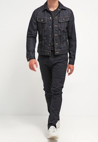 Dunkelblaue Jeansjacke mit zwei Brusttaschen und Metallknöpfen, kombiniert mit schwarzen Hosen und weißen Sneakers, im Slim-Fit-Design.