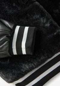 Svart fuskpäls med slät lämska. Ärmslutet har svarta och grå ränder, vilket ger kontrast till den strukturerade ytan.