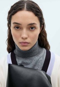 Svart läderkuvert som hålls mot en grå polotröja. Har ljusgrå och mörklila detaljer på ytterplagget. Neutral bakgrund.