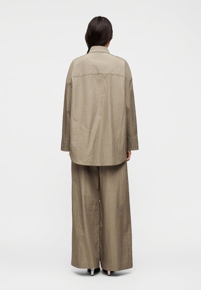 Γυναίκα με μακριά σκούρα μαλλιά, με την πλάτη γυρισμένη, φορώντας χαλαρό, υφασμάτινο μπεζ σακάκι και παντελόνι με φαρδιά πόδια, με μαύρα τακούνια, σε λευκό φόντο.
