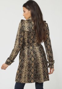 Abrigo trench de estampado de serpiente en beige y negro, con cintura ceñida con cinturón, mangas largas y cuello. Textura suave y diseño de longitud media.