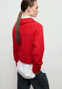Maglione rosso, corto, con un ampio colletto e polsini a coste, indossato sopra una camicia bianca. Lo sfondo è neutro, mettendo in risalto la texture del capo.