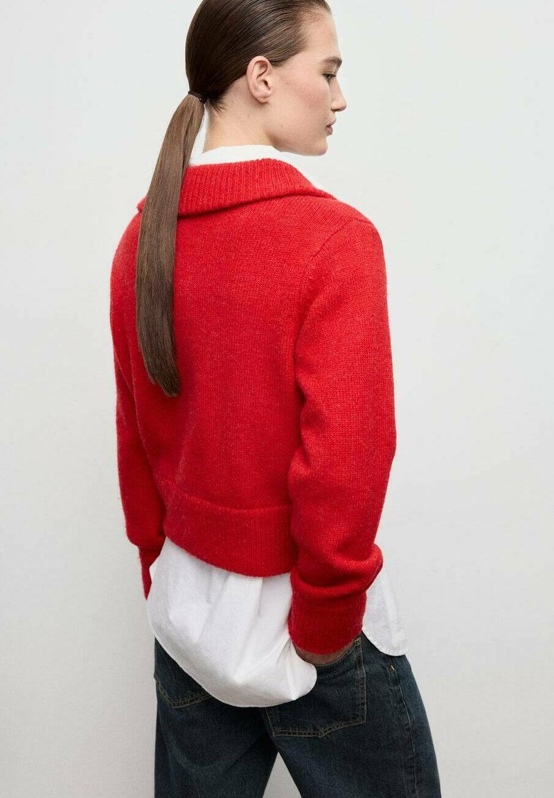 Maglione rosso, corto, con un ampio colletto e polsini a coste, indossato sopra una camicia bianca. Lo sfondo è neutro, mettendo in risalto la texture del capo.