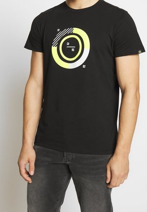 Homme portant un t-shirt noir avec un graphisme circulaire néon jaune et blanc et le texte « CA RS 1982 » sur le devant, associé à un jean gris foncé.