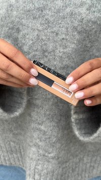 NARS concealer in een perzikachtige nude tint, in een rechthoekige tube met een zwarte applicator. Handen houden het product tegen een zachte grijze trui.