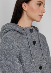 Manteau en mélange de laine gris avec une surface texturée, capuche et boutons noirs. Présente des accents métalliques sur les cordons de serrage et le col.