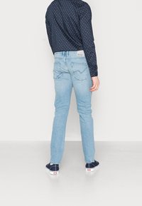 Person som bär ljusblå slim-fit jeans, marinblå skjorta med prickar och marinblå sneakers, stående mot en enkel vit bakgrund.