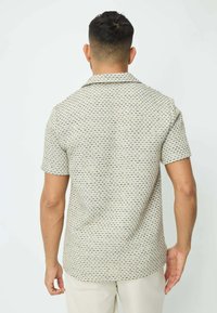 Chemise à manches courtes avec un fond crème clair, présentant un motif géométrique répétitif en bleu et noir, un col pointu et une coupe décontractée.