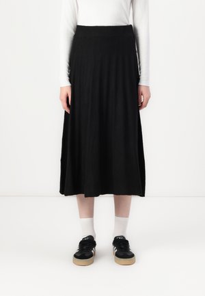 Personne portant une jupe midi noire, un haut à manches longues blanc, des chaussettes blanches et des baskets noires à semelles en gomme sur un fond blanc.