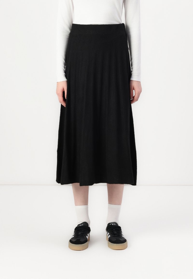 Personne portant une jupe midi noire, un haut à manches longues blanc, des chaussettes blanches et des baskets noires à semelles en gomme sur un fond blanc.