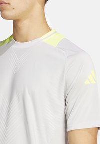 Lichtgrijs sportshirt met gele accenten. Bevat geometrische patronen en een ronde halsontwerp. Zachte textuur, korte mouwen.