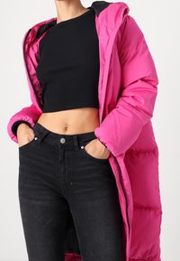 Abrigo acolchado largo fucsia con capucha, que presenta un cierre delantero con cremallera y puños elásticos, combinado con unos vaqueros negros de cintura alta.