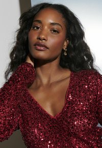 Haut en sequins rouges à manches longues et décolleté en V profond, présentant une texture lisse et des bords brodés, scintillant sous la lumière.