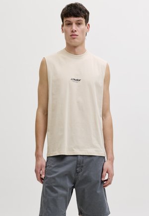 Jeune homme aux cheveux bruns courts portant un débardeur beige avec l'inscription « JJ Studios » et un short gris, debout devant un fond clair uni.
