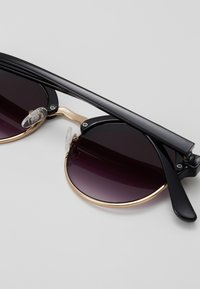 Gafas de sol negras con lentes de un púrpura oscuro degradado, acentos dorados y un puente de metal. Presentan almohadillas nasales transparentes y brazos gruesos negros.